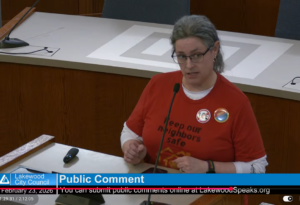 The image depicts an individual making public comment at Lakewood City Council, wearing a red shirt and anti-ICE pins