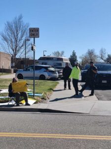 Woman sitting in chair holding sing reading "Come get this sign" while another woman talks to two laughing police officers